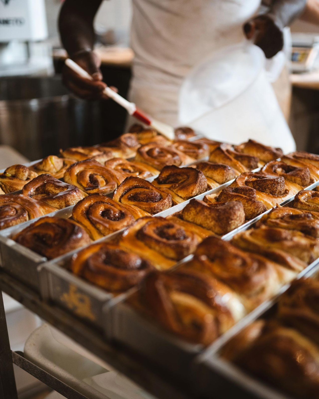Ein Bäcker bestreicht frisch gebackene Zimtschnecken in einer Metallform. Die Zimtschnecken sind goldbraun und gleichmäßig angeordnet.