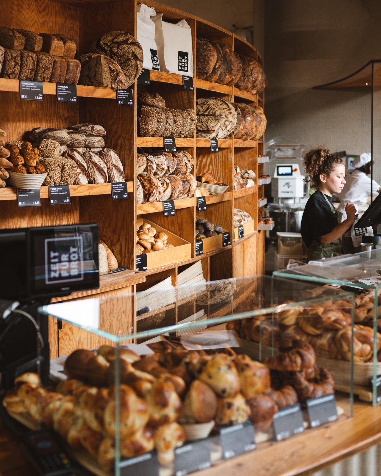 Holzregal mit verschiedenen Brotsorten und Brötchen. Im Vordergrund eine Glasvitrine mit frischen Backwaren.